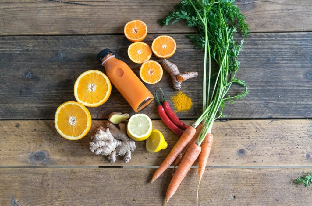 Oranges, carrots, ginger, lemons and chillies on a table beside a freshly pressed juice to indicate what is a cold press juicer